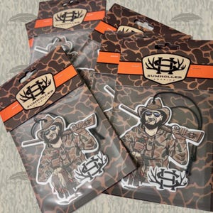 May include: Several air fresheners in clear plastic packaging. Each features a cartoon illustration of a man in a cowboy hat and camouflage clothing, holding a rifle and a duck. The packaging has a brown and orange design with the text "SUMHOLLER AMERICA".