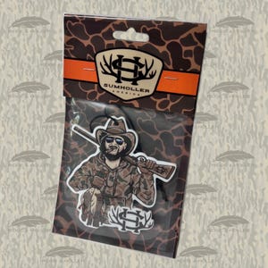 May include: An air freshener featuring a hunter in camouflage clothing, sunglasses, and a cowboy hat, holding a rifle and a duck. The packaging includes the text "SUMHOLLER AMERICA" and a logo with antlers. The background has a camouflage pattern.