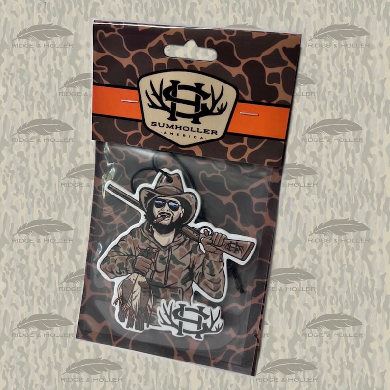 May include: An air freshener featuring a hunter in camouflage clothing, sunglasses, and a cowboy hat, holding a rifle and a duck. The packaging includes the text "SUMHOLLER AMERICA" and a logo with antlers. The background has a camouflage pattern.