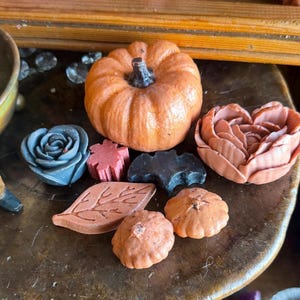 May include: A collection of wax figures including a large orange pumpkin, a dark gray rose, a small red star, a black bat, a peach-colored flower, a brown leaf-shaped piece, and two small orange pumpkins with textured surfaces.