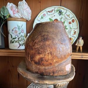 May include: A vintage, weathered wooden hat block with a wide base and a rounded top. The hat block sits on a carved wooden stand. A ceramic jar with floral illustrations and a decorative plate are in the background.