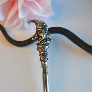 May include: A silver-toned metal hair stick featuring a detailed topper shaped like a crescent moon with a face wearing a jester-style cap. The long, slender, dark-colored shaft curves slightly, extending downward from the metallic moon ornament.
