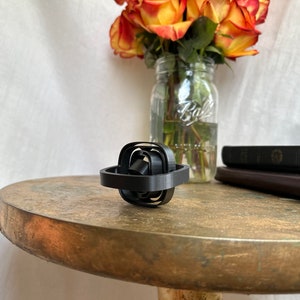 May include: A black, multi-layered, geometric, 3D printed object with a square shape. The object is sitting on a wooden table.