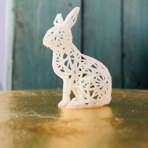 May include: A white, openwork rabbit figurine, showcasing a delicate, web-like design. The rabbit is posed in a seated position, with its ears erect. The figurine is set against a gold surface and a teal-colored backdrop.