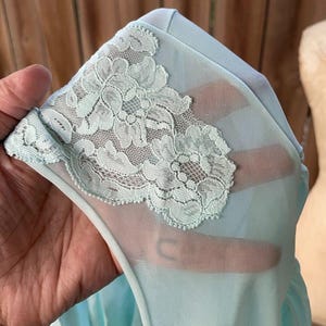 May include: Close-up of a light blue vintage nightgown. The sheer fabric is accented with delicate, pale green lace floral detailing along the neckline and shoulder. The nightgown is made of a soft, flowing material.