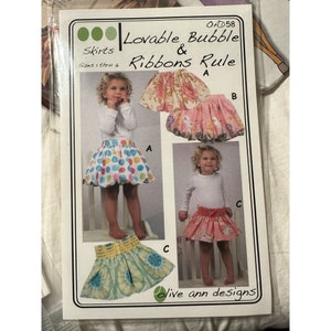 May include: A sewing pattern for making children's skirts. The pattern includes three different skirt styles: a bubble skirt with polka dots, a floral skirt, and a solid pink skirt. The pattern is called "Lovable Bubble & Ribbons Rule" and is designed by "olive ann designs".