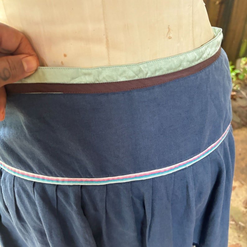 Puede incluir: Una falda azul con una cintura de cinta marr&oacute;n y azul claro y un ribete blanco, rosa y azul en el dobladillo.