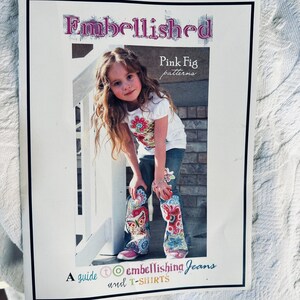 May include: A book cover with the title "Embellished" in purple script. The cover features a young girl wearing a white t-shirt and jeans with colorful floral embellishments. The text "Pink Fig patterns" is also visible.