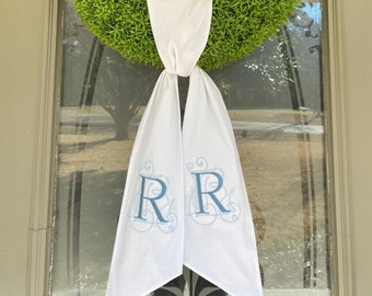 May include: A green wreath with a white ribbon with the letters "RR" embroidered in blue.