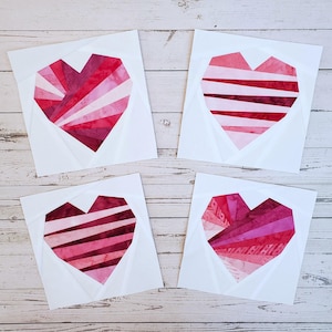 Könnte beinhalten: Vier quadratische Stoffblöcke mit geometrischen Herzmustern. Jedes Herz besteht aus Stoffen in Rot- und Rosatönen. Die Herzen sind vor einem weißen Hintergrund mit einem gefalteten Papiereffekt angeordnet. Die Blöcke liegen auf einer hellen Holzoberfläche.