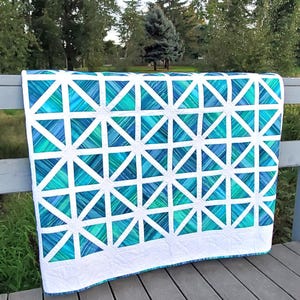 Puede incluir: Una colcha blanca con un patrón geométrico de triángulos azules y verdes. La colcha está colgada en una barandilla de madera.