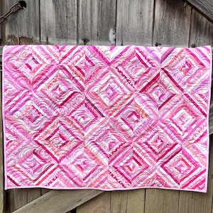 Puede incluir: Un edredón rosa y blanco con un patrón geométrico. El edredón presenta un diseño de diamantes repetidos con diferentes tonos de rosa y blanco. El edredón se muestra sobre un fondo de madera desgastada.