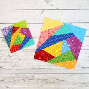 May include: Two colourful patchwork quilt blocks made from fabric scraps in various patterns and colours, including red, yellow, green, blue, and purple. The blocks are arranged on a white wooden surface.