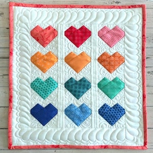 May include: A quilted wall hanging featuring nine colorful heart-shaped patches. The hearts are arranged in a 3x3 grid on a white background with a feather-like quilted pattern. The border is a coral pink with a floral pattern.
