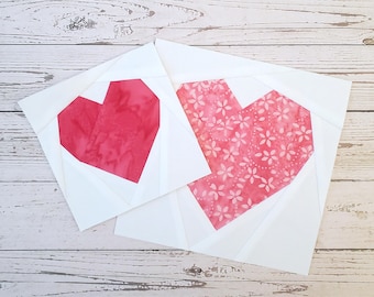 May include: Two square fabric quilt blocks featuring heart designs. One block displays a solid red heart, while the other features a larger heart with a pink and white floral pattern. Both hearts are centered on white fabric backgrounds.