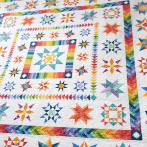 May include: A white quilt featuring a central star block surrounded by a rainbow-colored border of small triangles and various star-shaped patchwork patterns in shades of red, orange, yellow, green, blue, and purple with intricate quilting.