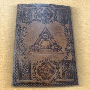 May include: A brown leather journal with an embossed design of an eye inside a triangle. The journal has a decorative border with intricate patterns.