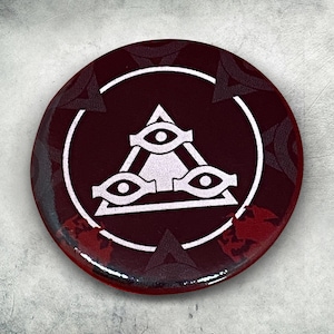 May include: A red button with a white triangle and three eyes inside. The button has a black and white pattern around the edge.