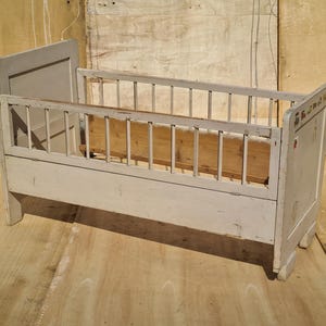 May include: A vintage, pale grey wooden cot with a slatted side and headboard. The cot has a wooden base and a light-coloured wooden interior. The cot is designed with a classic, vintage aesthetic.