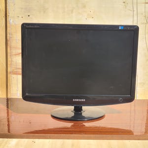 Könnte beinhalten: Ein schwarzer Samsung Computermonitor mit glänzendem Bildschirm und rundem Fuß. Der Monitor steht auf einer braunen Holzoberfläche. Der Modellname "SyncMaster 2032w" ist oben links aufgedruckt.