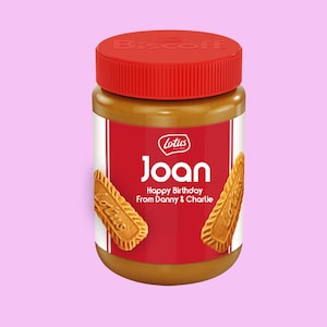 Peut inclure: Un pot de pâte à tartiner Lotus Biscoff avec un couvercle et une étiquette rouges. L'étiquette indique "Joan Happy Birthday From Danny & Charlie" avec deux biscuits Biscoff de chaque côté. Le pot est rempli d'une pâte à tartiner brun clair.