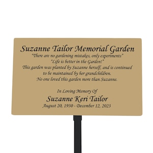 May include: A brown metal memorial garden sign with the text "Suzanne Tailor Memorial Garden" and the quote "There are no gardening mistakes, only experiments."  The sign also includes the text "In Loving Memory Of Suzanne Keri Tailor" and the dates "August 20, 1930 - December 12, 2023".