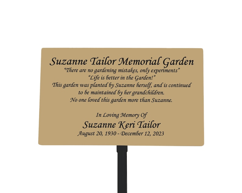 May include: A brown metal memorial garden sign with the text "Suzanne Tailor Memorial Garden" and the quote "There are no gardening mistakes, only experiments."  The sign also includes the text "In Loving Memory Of Suzanne Keri Tailor" and the dates "August 20, 1930 - December 12, 2023".