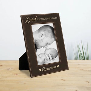 May include: Brown leather picture frame with a stitched border. The frame has a gold inscription that reads "Dad Established 2023" and "Cameron" with a heart symbol. A black and white photo of a father and baby is in the frame.