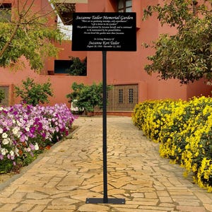May include: A black metal sign with a gold inscription in a garden setting. The sign reads "Suzanne Tailor Memorial Garden. There are no gardening mistakes, only experiments. Life is better in the garden. This garden was planted by Suzanne herself, and is continued to be loved by her grandchildren. No one is measured by their business more than by the love they leave behind. In loving memory of Suzanne Keri Tailor. August 28, 1970 - December 12, 2023."