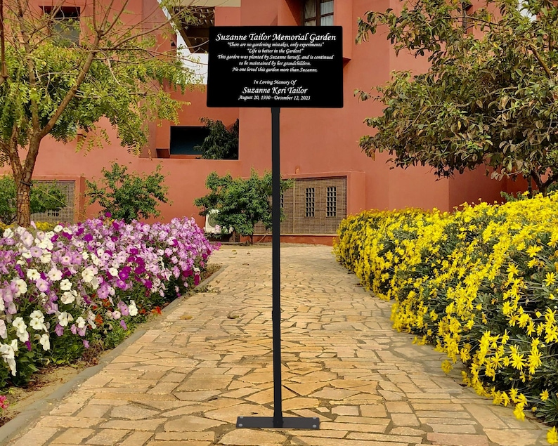 May include: A black metal sign with a gold inscription in a garden setting. The sign reads "Suzanne Tailor Memorial Garden. There are no gardening mistakes, only experiments. Life is better in the garden. This garden was planted by Suzanne herself, and is continued to be loved by her grandchildren. No one is measured by their business more than by the love they leave behind. In loving memory of Suzanne Keri Tailor. August 28, 1970 - December 12, 2023."