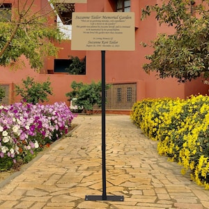 May include: A gold plaque with a black metal stand in a garden setting. The plaque reads "Suzanne Taylor Memorial Garden" and includes a quote about the garden. The plaque also states "In loving memory of Suzanne Kerr Taylor August 20, 1948 - December 12, 2023."