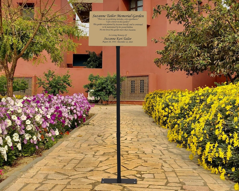 May include: A gold plaque with a black metal stand in a garden setting. The plaque reads "Suzanne Taylor Memorial Garden" and includes a quote about the garden. The plaque also states "In loving memory of Suzanne Kerr Taylor August 20, 1948 - December 12, 2023."