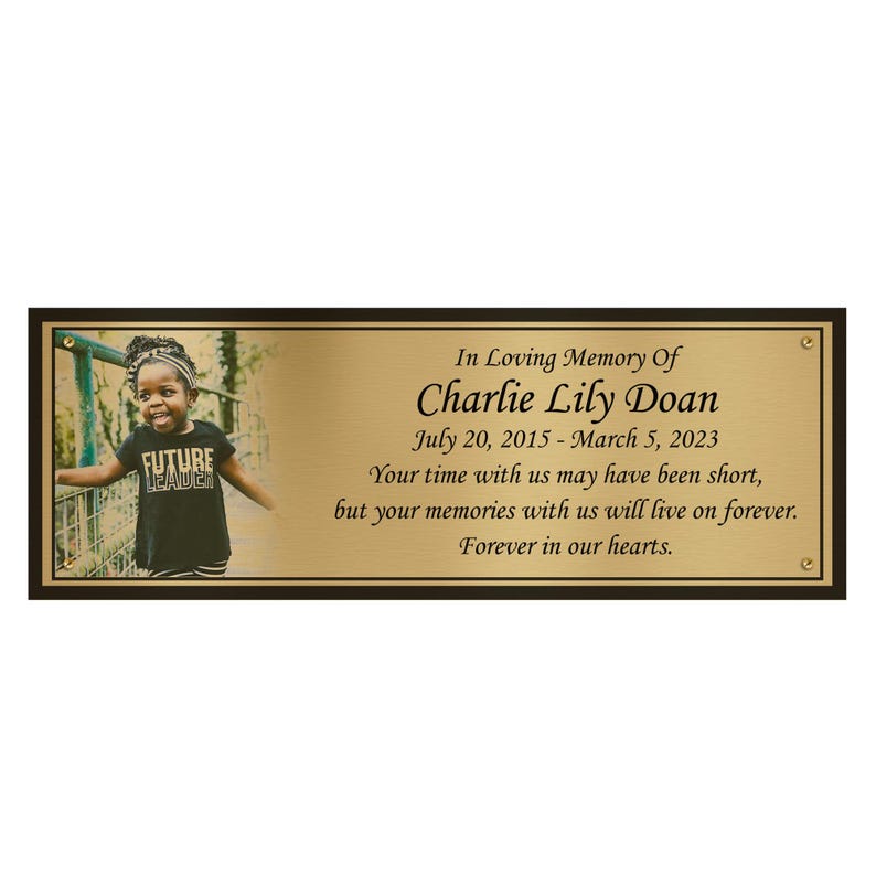 Pode incluir: Uma placa memorial dourada com uma foto de uma menina pequena vestindo uma camiseta preta com as palavras "Future Leader". A placa diz "In Loving Memory Of Charlie Lily Doan 20 de julho de 2015 - 5 de mar&ccedil;o de 2023 Seu tempo conosco pode ter sido curto, mas suas mem&oacute;rias conosco viver&atilde;o para sempre. Para sempre em nossos cora&ccedil;&otilde;es."