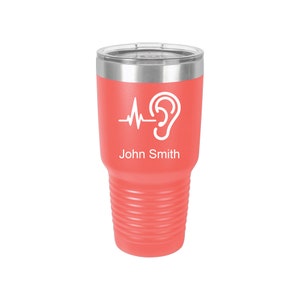 May include: Coral pink stainless steel tumbler with a white ear and heartbeat graphic and the name "John Smith" printed on the side.