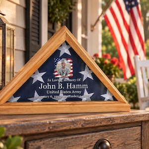 Puede incluir: Una vitrina triangular de madera que contiene una bandera plegada con un patrón de estrellas. El centro presenta una fotografía cuadrada de un soldado en uniforme, con el texto: In Loving Memory Of John B. Hamm, United States Army, January 6, 1995 - December 2, 2018.
