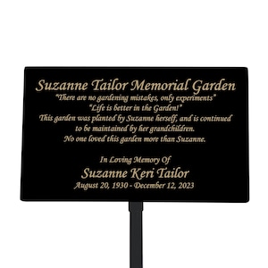 May include: Black metal memorial garden sign with gold lettering. The sign reads "Suzanne Tailor Memorial Garden" and includes a quote about gardening. The sign also includes the dates "August 20, 1930 - December 12, 2023".