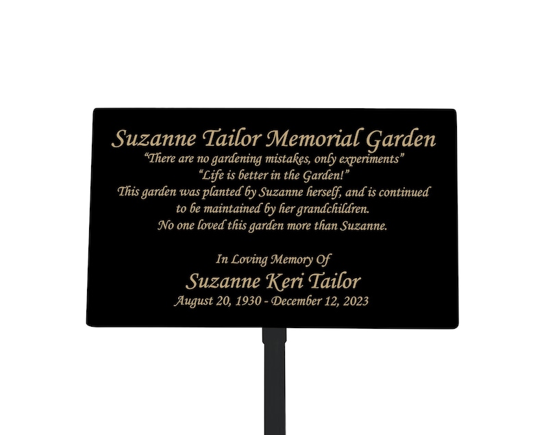 May include: Black metal memorial garden sign with gold lettering. The sign reads "Suzanne Tailor Memorial Garden" and includes a quote about gardening. The sign also includes the dates "August 20, 1930 - December 12, 2023".