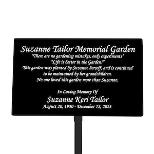 May include: Black metal memorial garden sign with white text. The sign reads "Suzanne Tailor Memorial Garden" and includes a quote about gardening. The sign also includes the name "Suzanne Keri Tailor" and dates "August 20, 1930 - December 12, 2023".
