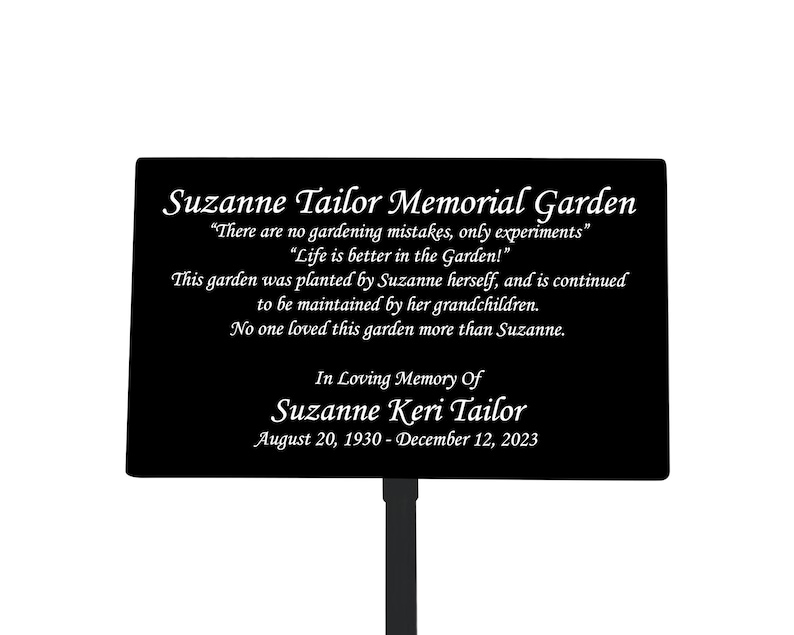 May include: Black metal memorial garden sign with white text. The sign reads "Suzanne Tailor Memorial Garden" and includes a quote about gardening. The sign also includes the name "Suzanne Keri Tailor" and dates "August 20, 1930 - December 12, 2023".