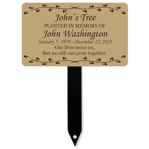 Op de afbeelding: Een bruine metalen tuinpaal met een zwarte metalen paal. Het bordje staat er op: "John's Tree Geplant ter nagedachtenis aan John Washington 5 januari 1970 - 22 december 2023 Ons leven gaat verder, Maar we kunnen nog steeds samen groeien."