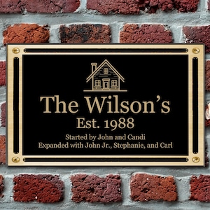 May include: A black plaque with gold trim and screws, mounted on a brick wall. The plaque features a house icon, the text "The Wilson's Est. 1988", and the text "Started by John and Candi Expanded with John Jr., Stephanie, and Carl".