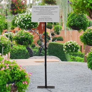 May include: A black metal sign holder with a white sign that reads "Suzanne Taylor Memorial Garden. There are no gardening mistakes, only opportunities to learn. Life is better in the garden. This garden was planted by Suzanne herself and is continued to be maintained by her family and friends. No one loved this garden more than Suzanne. In loving memory of Suzanne Kerr Taylor August 20, 1970 - December 12, 2021."