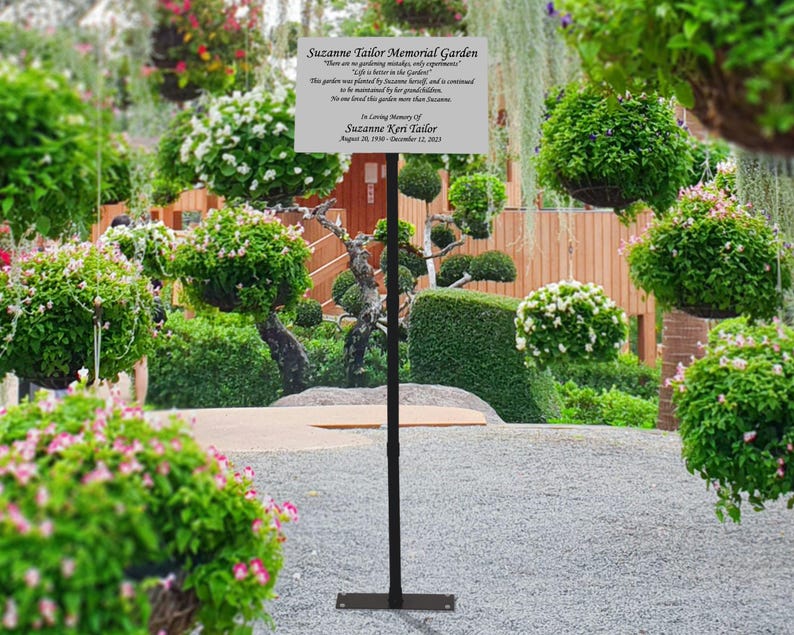 May include: A black metal sign holder with a white sign that reads "Suzanne Taylor Memorial Garden. There are no gardening mistakes, only opportunities to learn. Life is better in the garden. This garden was planted by Suzanne herself and is continued to be maintained by her family and friends. No one loved this garden more than Suzanne. In loving memory of Suzanne Kerr Taylor August 20, 1970 - December 12, 2021."