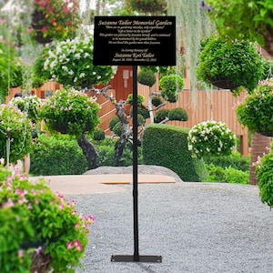 May include: A black metal sign with gold lettering in a garden setting. The sign reads "Suzanne Tailor Memorial Garden.  There are no gardening boundaries, only opportunities.  Life is better in the garden.  This garden was planted by Suzanne and is continued to be maintained by her family.  We are blessed by her presence.  In Loving Memory of Suzanne Kerr Tailor.  August 20, 1958 - December 12, 2021."