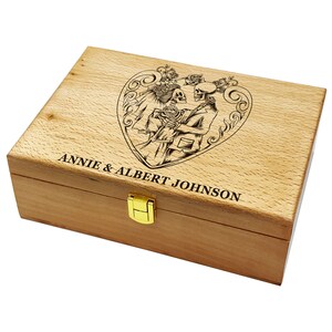 Peut inclure: Une boîte en bois avec un fermoir doré. Le couvercle est gravé d'un motif de cœur représentant deux squelettes se tenant la main. Le texte "ANNIE & ALBERT JOHNSON" est gravé sous le cœur.