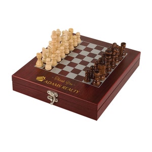 May include: A mahogany wood chess set with a hinged lid and a gray and white chessboard. The chess set is closed and the lid has the text "Thank You - ADAMS REALTY" in gold lettering.