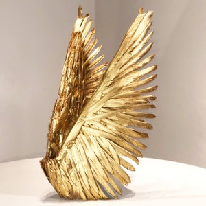 Puede incluir: Una escultura dorada de una sola ala, con plumas detalladas. El ala se exhibe sobre un fondo neutro, resaltando su brillo metálico y su intrincada textura. La forma del ala sugiere un tema de vuelo o libertad.