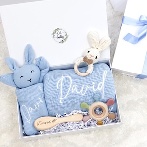 May include: A baby gift set in a white box, featuring a blue blanket with the name "David", a blue bunny plush, a wooden brush, and two wooden toys. The box is tied with a light blue ribbon.