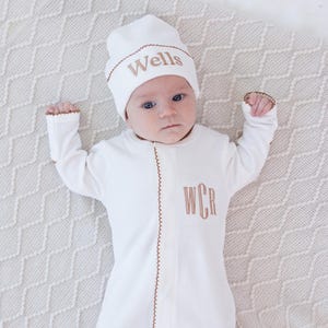 May include: A white baby sleeper and matching hat with brown trim and embroidered text. The sleeper has the initials "WCR" embroidered on the front. The hat is embroidered with the name "Wells". The baby is lying on a cream-colored blanket.