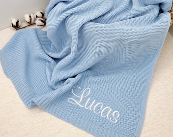 May include: A light blue knitted baby blanket with the name "Lucas" embroidered in white script. The blanket is folded and draped, showing the texture of the knit. The blanket is displayed on a white surface.
